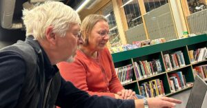 To personer sitter ved en pc i et rom med bøker (bibliotek)
